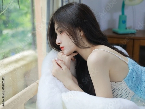 Fototapeta Woman in a blue crochet top resting by the window with a serene expression.