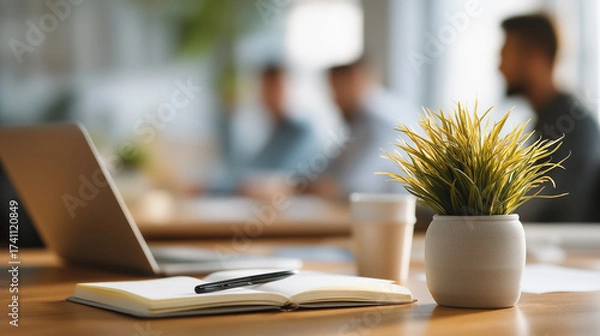 Fototapeta A plant, notebook, and pen with coworkers using laptops