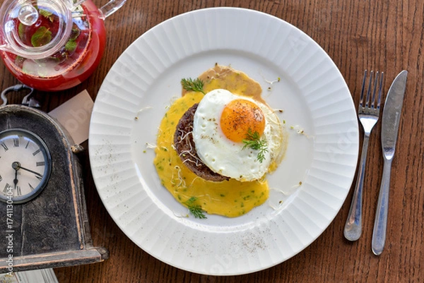 Fototapeta steak with sauce topped with fried egg on a plate on a table with tea and a retro clock