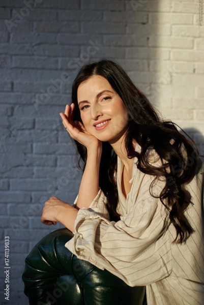 Fototapeta woman with long dark hair is sitting against a white brick wall, smiling gently while resting her chin on her hand in a warm, inviting atmosphere