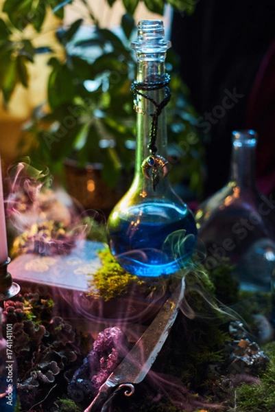 Fototapeta vibrant blue potion sits in a glass bottle, surrounded by green plants and soft candlelight. Smoke gently swirls around a mystical scene, creating an enchanting atmosphere