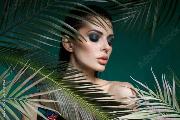 Fototapeta model showcases stunning makeup with green tones while posing amid lush tropical leaves. scene is full of vibrant colors, creating an eye-catching look