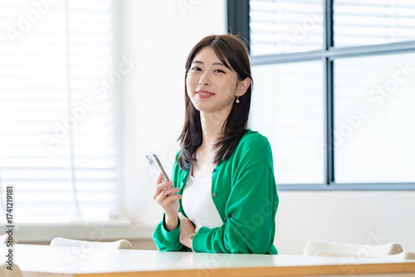 Fototapeta 明るい室内でスマートフォンを持ってカメラ目線で微笑む女性