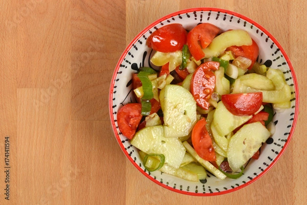 Fototapeta An overhead shot features a bowl of colorful salad made with sliced tomatoes, cucumbers, peppers, and spring onions, set against a light wooden background