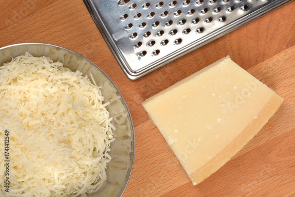 Fototapeta An overhead shot shows a wedge of Parmesan cheese, a pile of grated cheese in a metal bowl, and a cheese grater on a wooden cutting board