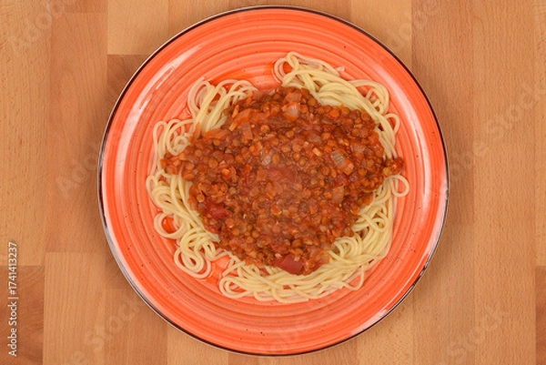 Fototapeta An overhead shot of a plate of spaghetti topped with thick, rich vegan lentil Bolognese sauce, served on an orange dish on a wooden background