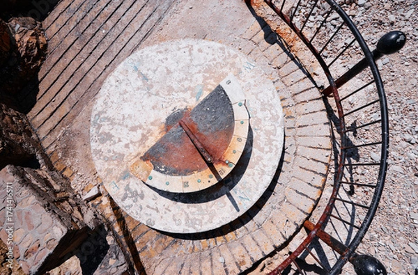 Fototapeta Aerial view of a weathered sundial with a rusty gnomon, surrounded by a metal railing on a stone platform.