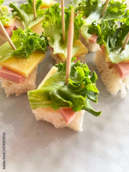 Fototapeta Small canapés made from square pieces of bread, salami, hard cheese, and fresh lettuce, held together with a wooden toothpick, are arranged on a gray textured plate. Close-up. Appetizer. Buffet.