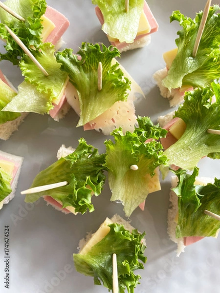 Fototapeta Small canapés made from square pieces of bread, salami, hard cheese, and fresh lettuce held together with a wooden toothpick are arranged on a gray textured plate, close-up, top view. Appetizer.