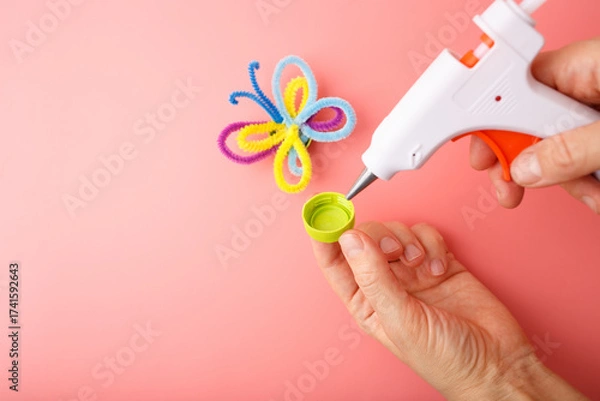 Fototapeta Crafting moment with hot glue gun and colorful pipe cleaner butterfly on pink background, ideal for educational content, DIY kits, kids activity brands, creative learning visuals,