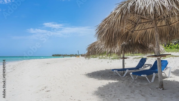 Fototapeta Two sun loungers on the sandy beach in the shade of straw umbrellas. Calm aquamarine ocean, blue sky, clouds. Cuba. Varadero. Resort. Hotel.