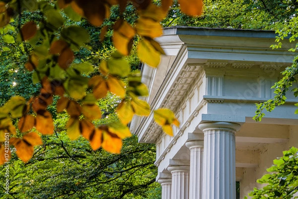 Fototapeta A classic pavilion with white columns in a green park, framed by autumn-colored foliage, illuminated by warm sunlight