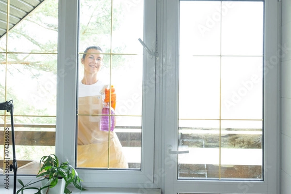 Fototapeta Woman in rubber gloves and apron manually washes window balcony of house with rag cleaner and mop outside behind glass. Restoring order and cleanliness in spring, cleaning servise