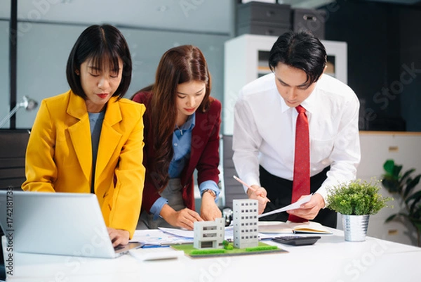Fototapeta Asian team collaborating on real estate and architecture projects with blueprints, models, and data charts in a modern office, showcasing business teamwork and success.