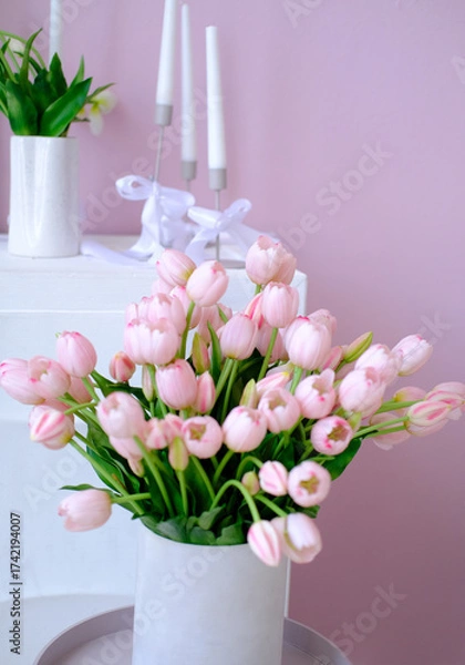 Fototapeta Pink tulips arranged in a white vase on a minimalist table, surrounded by elegant candles and greenery, creating a serene and inviting atmosphere for home decor