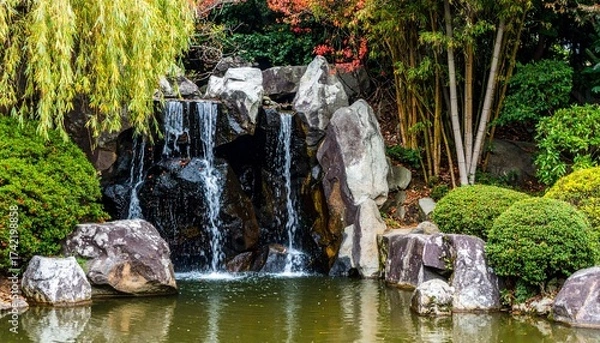 Fototapeta A gentle waterfall flows into a pond, framed by autumn foliage and lush garden greenery.