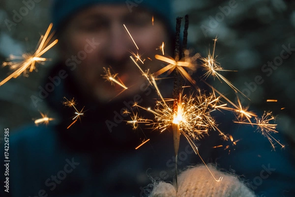 Fototapeta man wearing warm winter clothing holds sparkler, making bright sparks in cold outdoor setting. background is out focus, highlighting festive moment during winter gathering. close up.