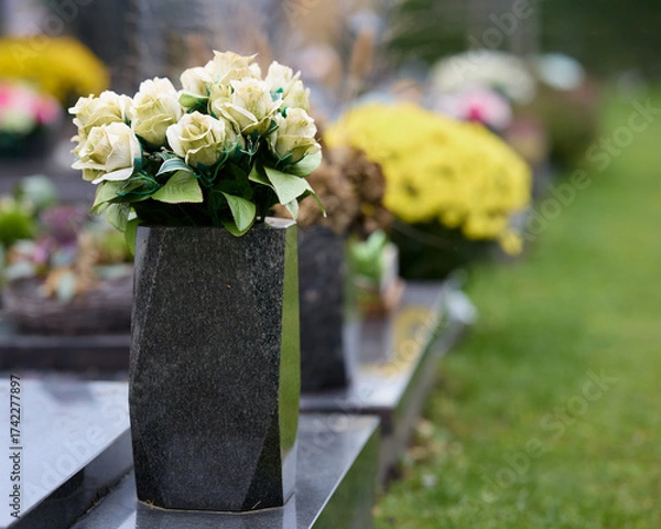 Fototapeta Elegant white roses in a dark stone vase adorn a headstone, set amidst other memorials and vibrant flowers in a serene, green cemetery landscape.