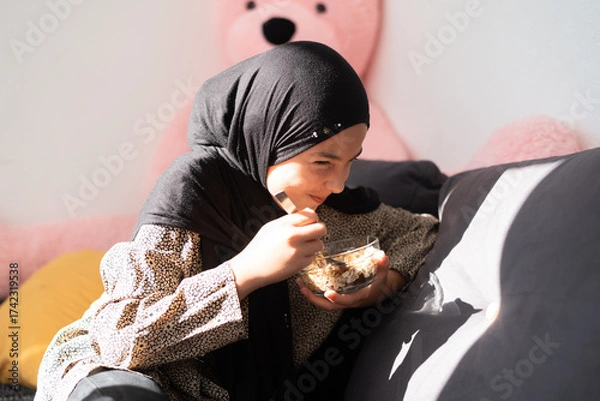 Fototapeta Young happy girl in hijab sitting on sofa eating a cereal meal, cozy home setting with soft sunlight and teddy bear in background.
