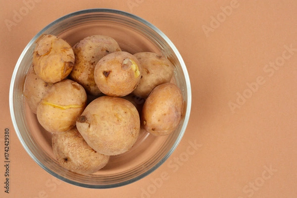 Fototapeta A glass bowl is filled with whole, boiled new potatoes, partially resting on a solid brown background with ample copy space