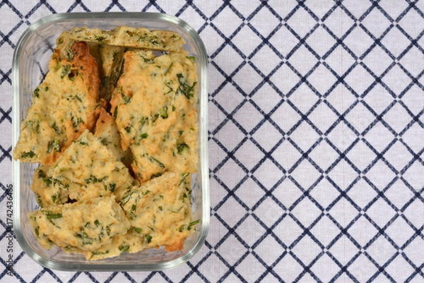 Fototapeta An overhead shot shows a glass container filled with homemade savory crackers with parsley, set against a blue and white checkered cloth