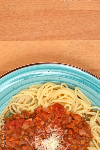 Fototapeta An overhead shot of a plate of spaghetti topped with vegan lentil Bolognese sauce and grated cheese, set on a blue dish over a wooden table