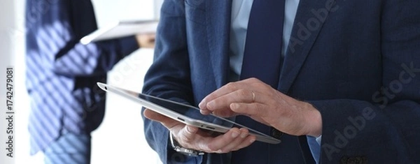 Fototapeta Businessman in a suit using a tablet during a corporate meeting, interacting with the touchscreen as blurred colleagues collaborate in the background, modern workplace tech
