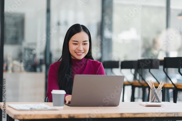 Fototapeta Young woman working on laptop in modern cafe, smiling while typing and enjoying coffee and creative workspace