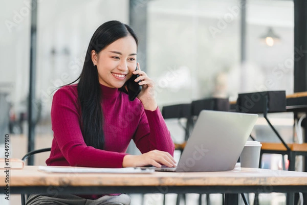 Fototapeta Young woman in red sweater talking on phone and working on laptop with coffee cup, smiling confidently in modern cafe