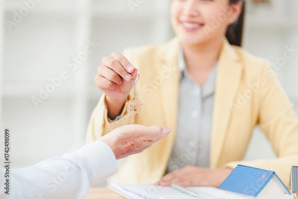 Fototapeta Real estate agent handing house key to client, smiling woman completing property sale with small model house on desk