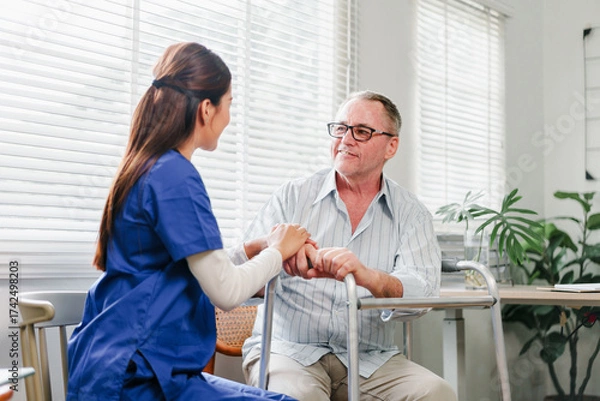 Fototapeta Friendly caregiver holding hands with senior man using walker in sunlit room, warm supportive interaction and calm atmosphere