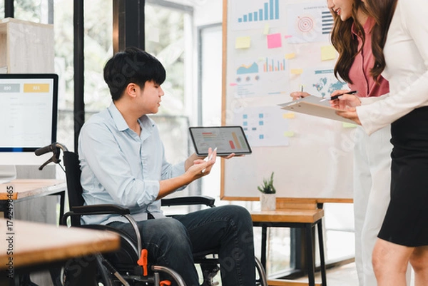 Fototapeta Young businessman in wheelchair presenting tablet with charts to colleagues, collaborative office meeting with charts and sticky notes