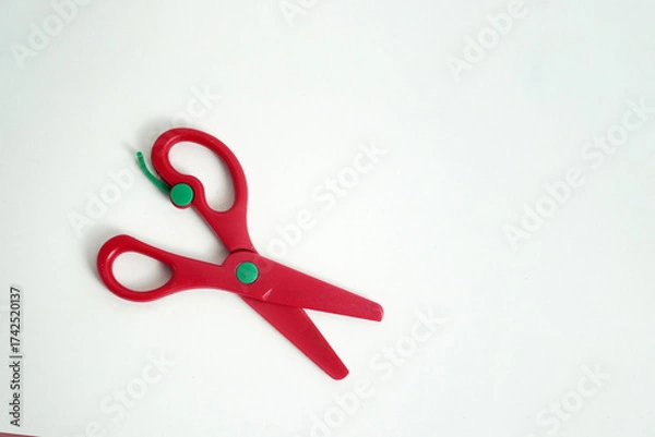 Fototapeta A pair of red plastic safety scissors with green pivot points, lying open on a clean white background. safe scissors for children learning to cut. school, crafts, art projects, and office supplies.