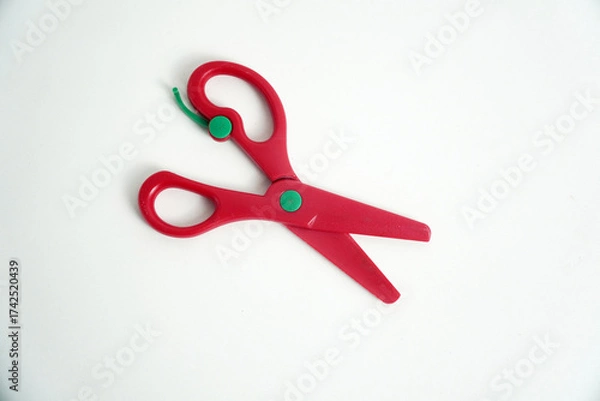 Fototapeta A pair of red plastic safety scissors with green pivot points, lying open on a clean white background. safe scissors for children learning to cut. school, crafts, art projects, and office supplies.