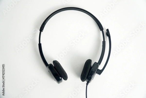 Fototapeta A pair of black on-ear headphones with a microphone boom, viewed from the top down on a white background. This image is perfect for concepts of a call center, customer service, communication, or music
