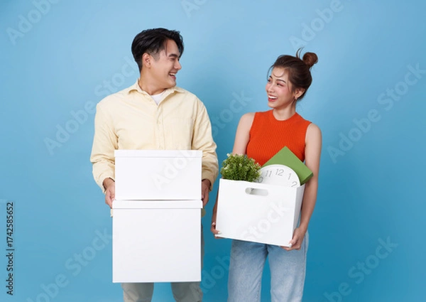 Fototapeta Asian couple carrying boxes and smiling isolated on blue background representing moving to new home teamwork lifestyle change and happiness in starting a new life together.