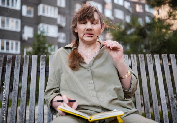 Fototapeta Elderly woman draws in notebook, pursues hobby, sitting on bench in city park. Active age, interesting life and creative abilities of older people