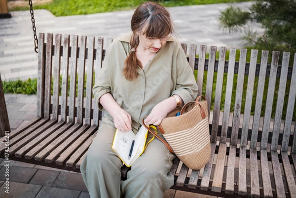Fototapeta Elderly woman draws in notebook, pursues hobby, sitting on bench in city park. Active age, interesting life and creative abilities of older people