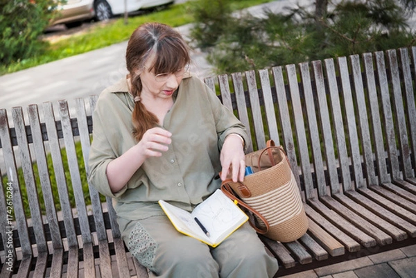 Fototapeta Elderly woman draws in notebook, pursues hobby, sitting on bench in city park. Active age, interesting life and creative abilities of older people