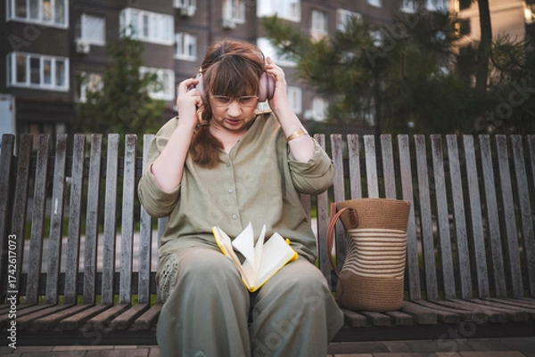 Fototapeta Aged woman in headphones draws in notebook, pursues hobby, sitting on bench in city park. Active age, interesting life and creative abilities of older people