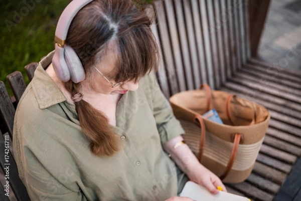 Fototapeta Aged woman in headphones draws in notebook, pursues hobby, sitting on bench in city park. Active age, interesting life and creative abilities of older people