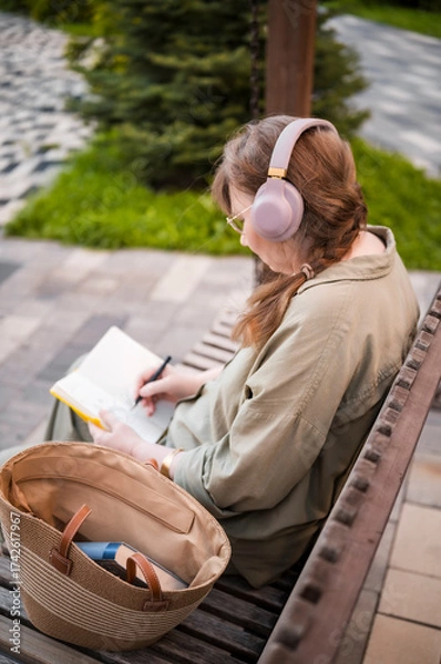 Fototapeta Elderly woman in headphones draws in notebook, pursues hobby, sitting on bench in city park. Active age, interesting life and creative abilities of older people