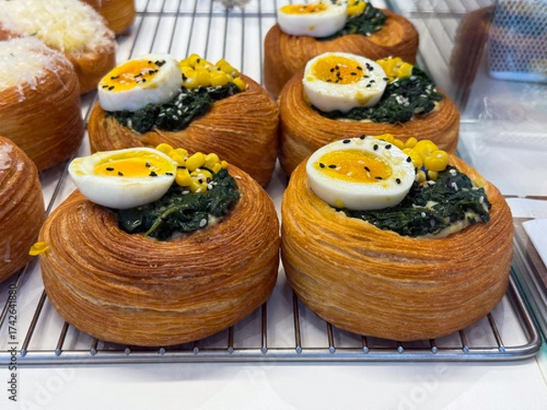 Fototapeta Fresh bakery pastries with egg, spinach, and corn topping on rack. Food photography, bakery products, and gourmet bread display.