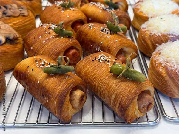 Fototapeta Fresh baked pastry rolls with sausage filling and pickled cucumber topping. Bakery products, gourmet snack, and culinary presentation on rack.