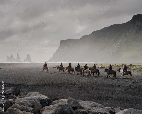 Fototapeta Group of horseback riders riding along a black sand beach in Iceland, surrounded by dramatic cliffs and misty coastal scenery. The moody atmosphere and natural setting create a cinematic, adventurous 