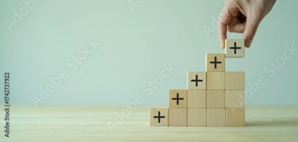 Fototapeta Building Positive: A hand delicately places a wooden block adorned with a plus sign atop a staircase, symbolizing growth, progress, and the addition of positive elements.