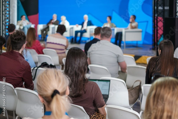 Fototapeta Panel discussion at modern conference featuring diverse speakers addressing audience against bright blue stage backdrop, capturing focus and professional energy