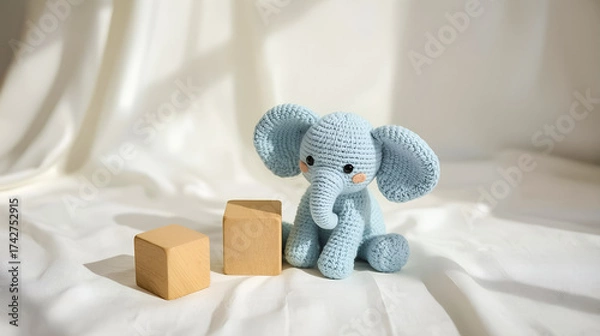 Fototapeta Charming blue handcrafted knitted elephant toy placed alongside wooden blocks on a sunny white fabric, evoking a sense of warmth and creativity.