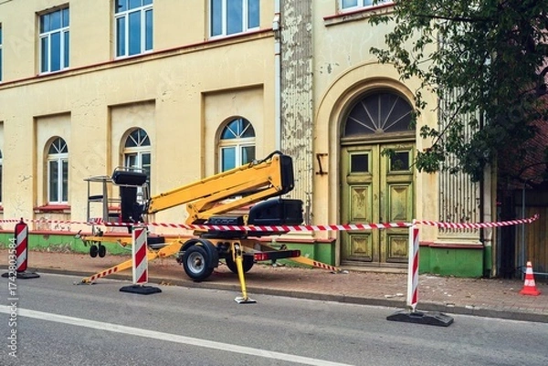 Fototapeta Construction site with a yellow mobile lift near an old building under renovation. The area is closed with red and white safety tape, indicating ongoing repair work and facade restoration.