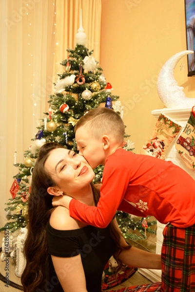 Obraz A woman and a boy sitting on the floor in front of a decorated Christmas tree. Garlands, ornaments, and baubles glitter on the tree. The comfort and joy of a family holiday